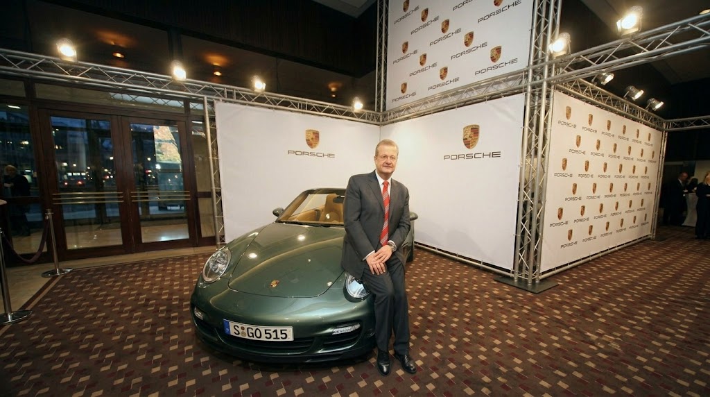 Former Porsche CEO Wendelin Wiedeking sits on the hood of a classic, vintage green Porsche 911 at a corporate press event in front of a branded backdrop featuring PORSCHE logos.